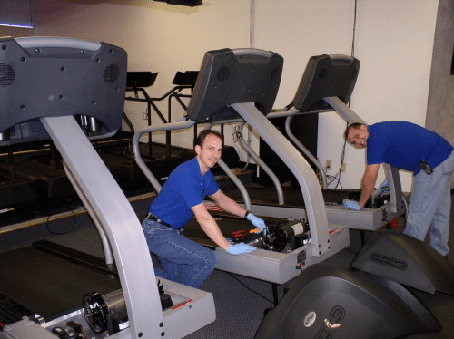 Two men repairing treadmills in a gym.
